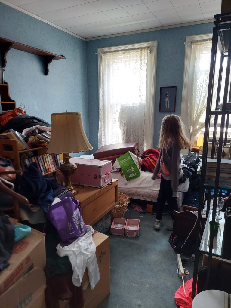 a smallish room with blue wallpaper, and full of piles of stuff. A young girl with her back to the camera almost disappears into the mess