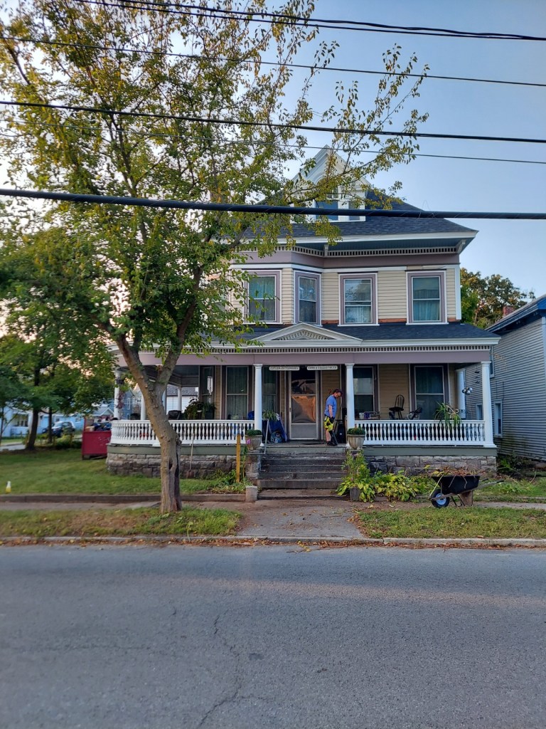 a large beige Victorian house with purplish trim, a full porch and bay window. There is a medium sized tree on the left, and a man standing on the top step holding a blower. There is clutter on the porch. There is unkempt landscaping on the right, the left is trimmed and there is a wheelbarrow on the side walk